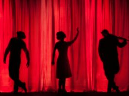 A dimly lit theatre with black silhouettes of performers against a red curtain.