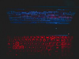 A laptop in a dark room. The screen has blue coding on it, and the keyboard is lit up in red.
