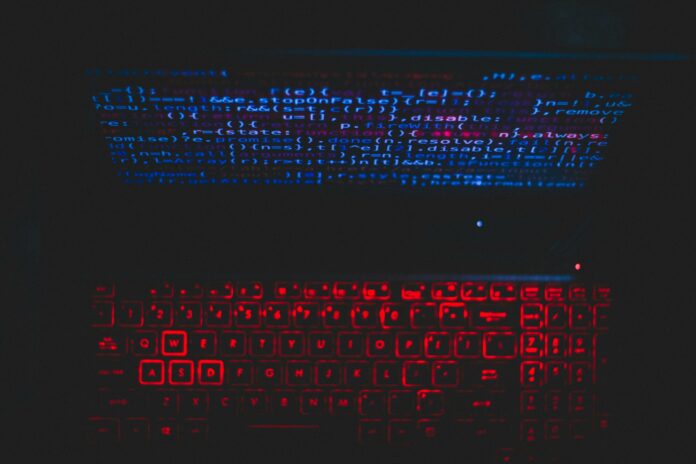 A laptop in a dark room. The screen has blue coding on it, and the keyboard is lit up in red.