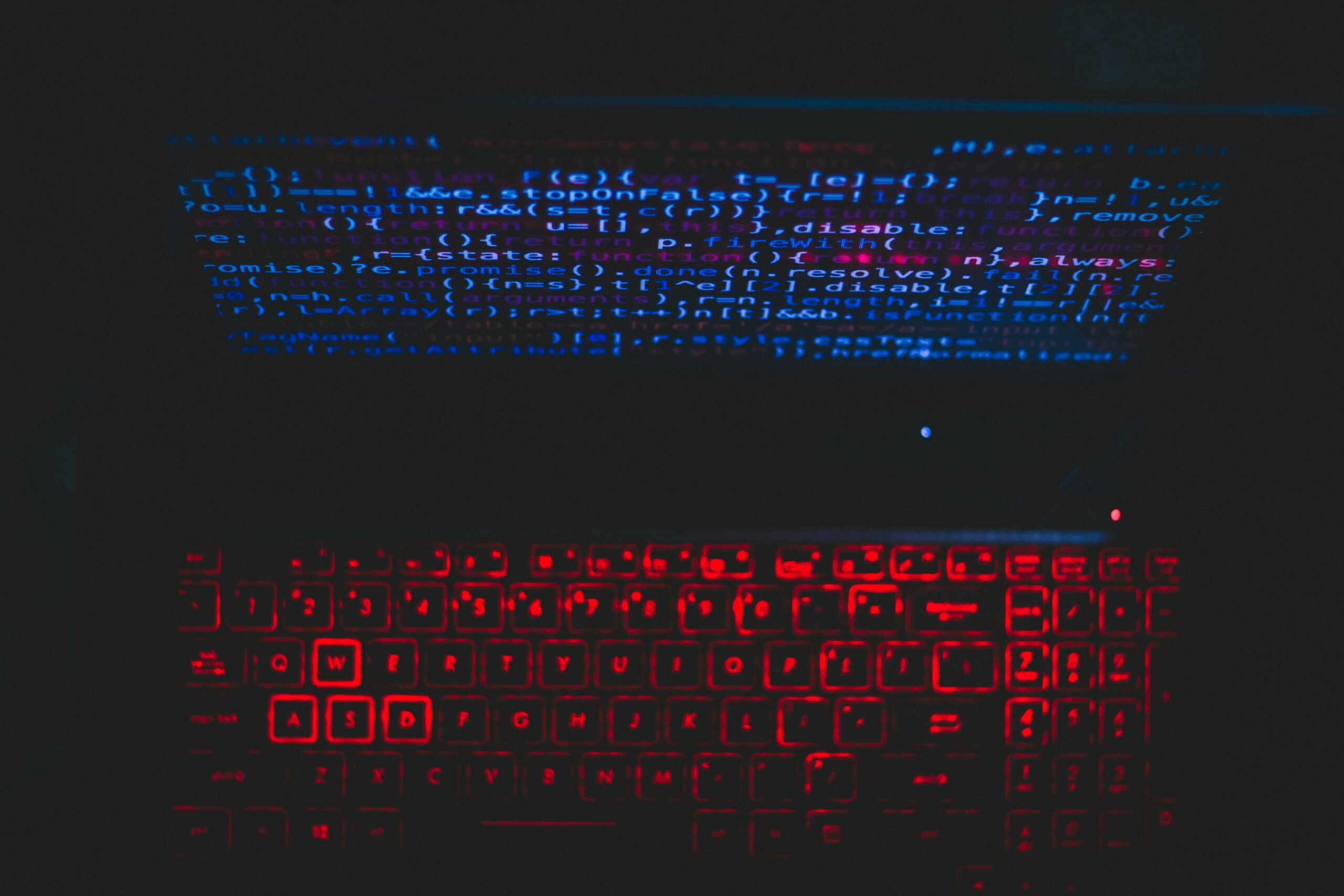 A laptop in a dark room. The screen has blue coding on it, and the keyboard is lit up in red.