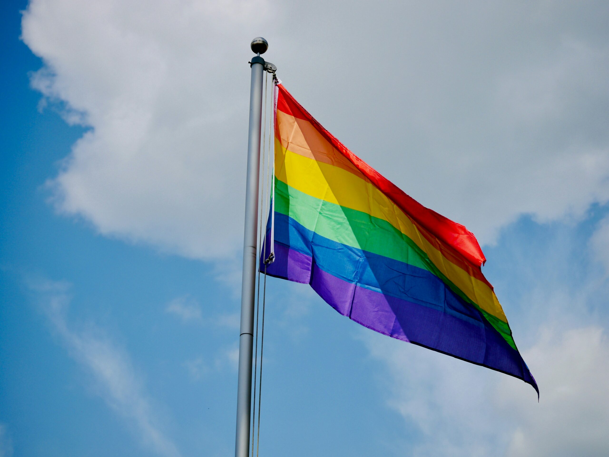 This is a photo of a pride flag, on a flag pole, blowing in the sky.