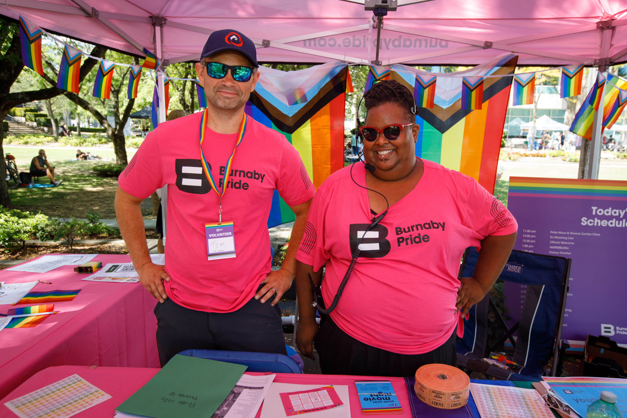 This is a photo of Athena Affan and another Burnaby Pride organizer. They are standing by a table, surrounded by pride flags, wearing “Burnaby Pride” t-shirts.