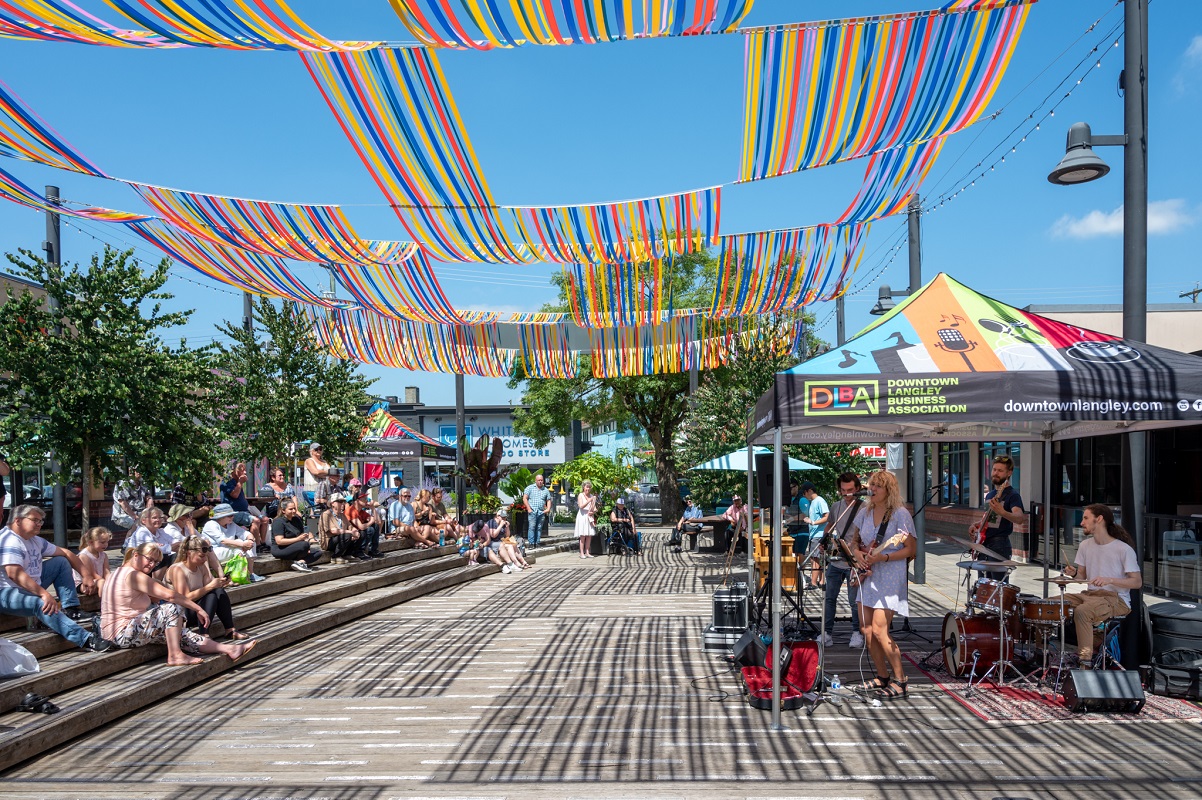 A large outdoor space with blue, yellow, and red streamers strung above it. A band made up of four individuals plays under a colourful tent while individuals and families watch while sitting on the wooden stairs of the outdoor space.