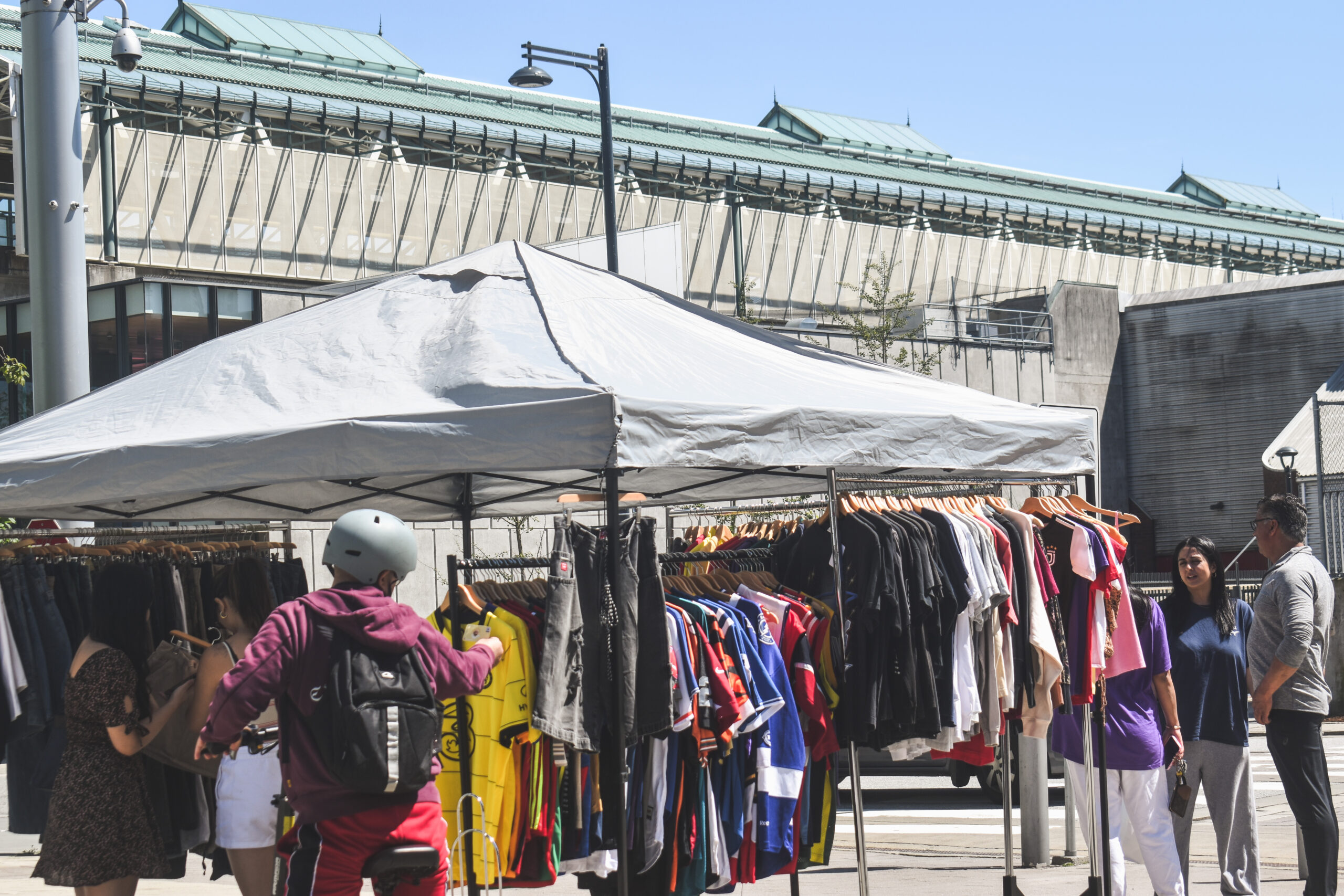 A rack of colourful clothing perched under a tent in front of a skytrain station.