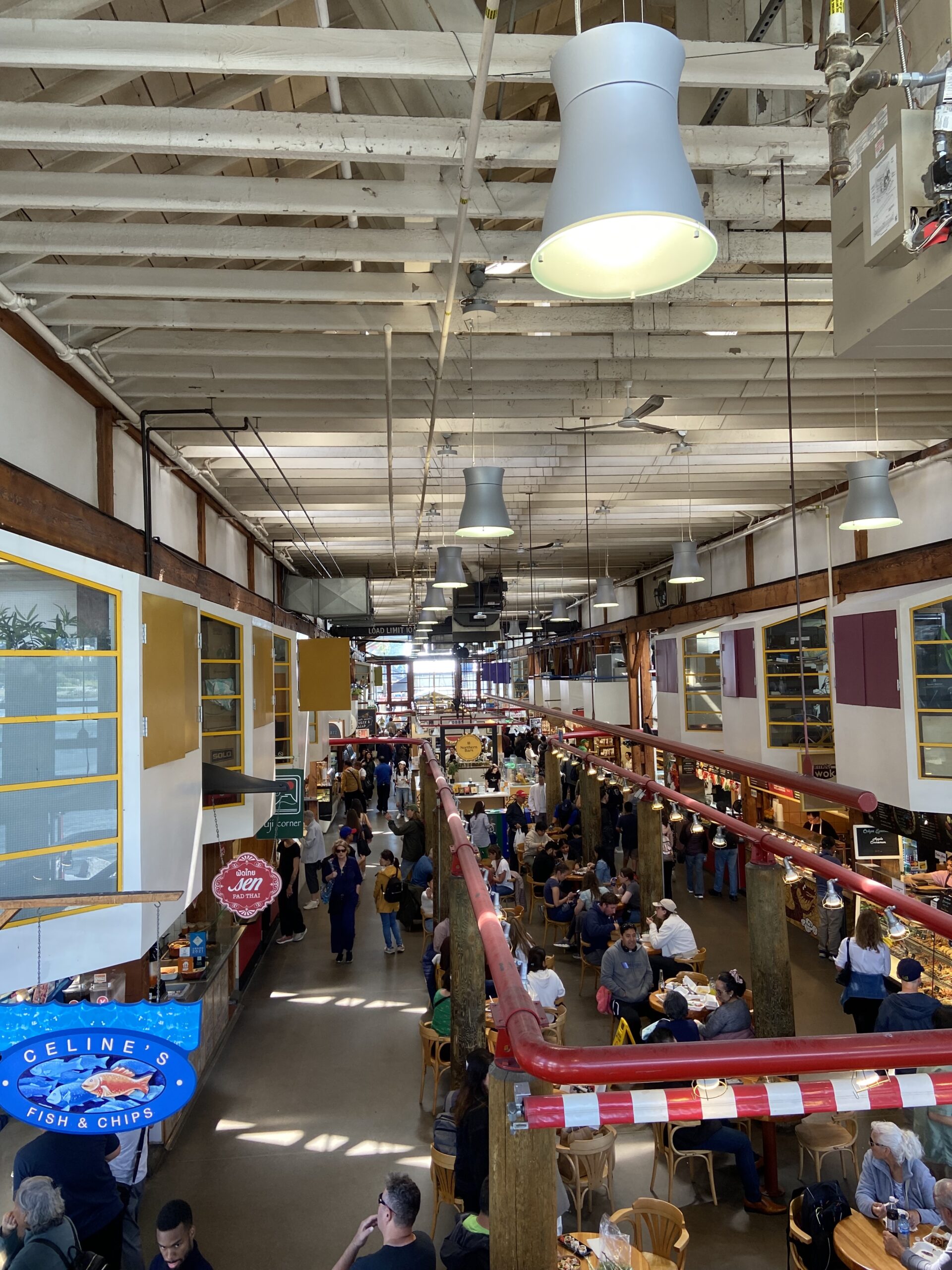 A top view of the food section of Granville Island Public Market. Various individuals sit at tables while enjoying food from the stalls surrounding them. Colorful awnings and signage are displayed throughout the space.