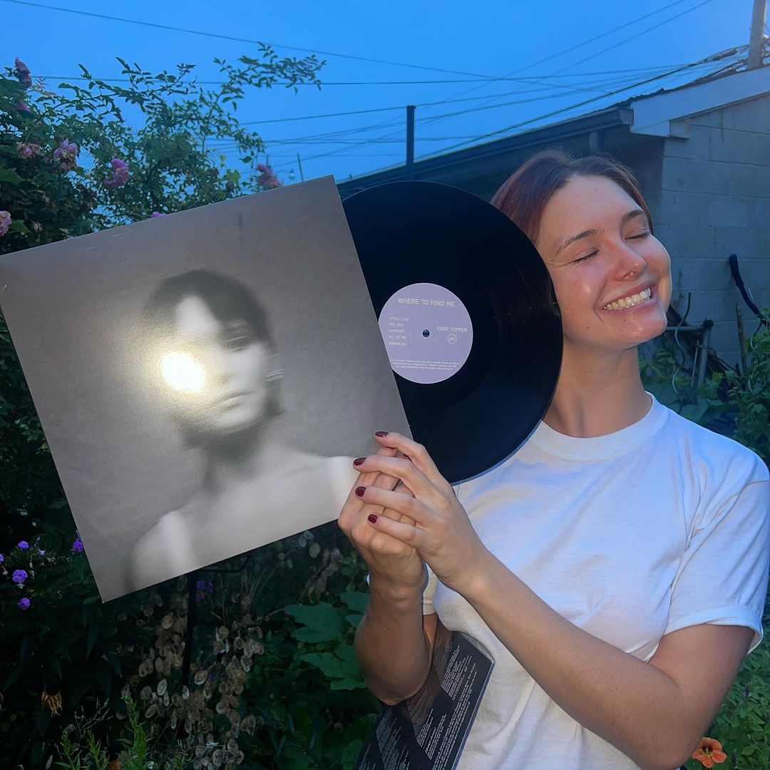 A girl with brown hair holding a vinyl next to her face. On the cover of the vinyl is a blurry gray photo of an individual with short hair.