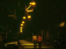 A grainy shot of two people walking down a street at night holding hands. The street is lit up by soft lights and decorated with cars.