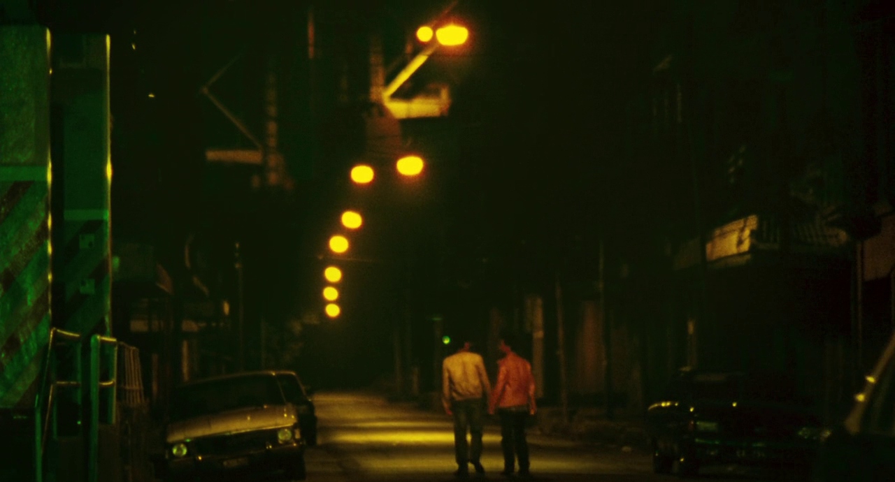 A grainy shot of two people walking down a street at night holding hands. The street is lit up by soft lights and decorated with cars.