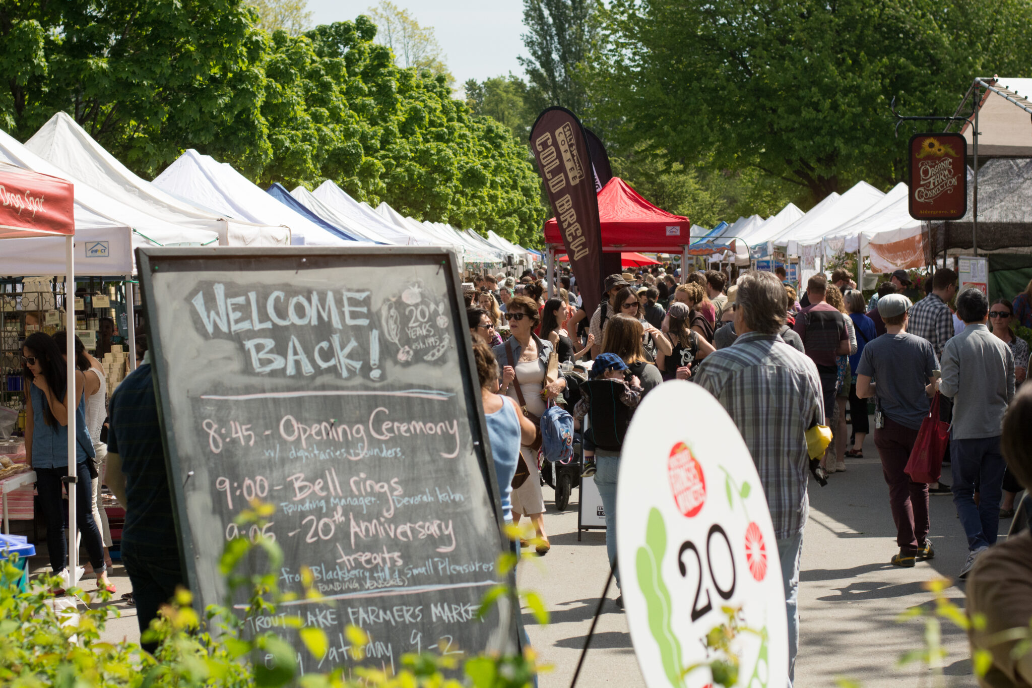 Crowded stalls surrounded by people and trees carrying various tiny trinkets and produce. A chalkboard sign reads “welcome back!” in pastel blue.