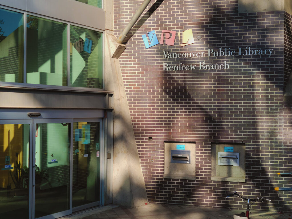 A brown brick building with book deposits and a motion-sensored glass door. The letters V, P, and L are written on the wall in blue, red, and yellow boxes respectively.