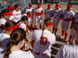 A group of girls huddle in a circle, each wearing a white uniform with red pinstripes and a red visor with a maple leaf on it.