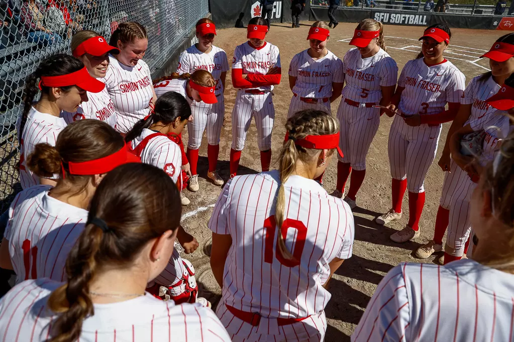 A group of girls huddle in a circle, each wearing a white uniform with red pinstripes and a red visor with a maple leaf on it.