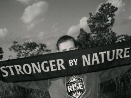 In black and white, Canadian soccer legend Christine Sinclair stands with an outstretched scarf covering her face. The scarf reads ‘stronger by nature.’