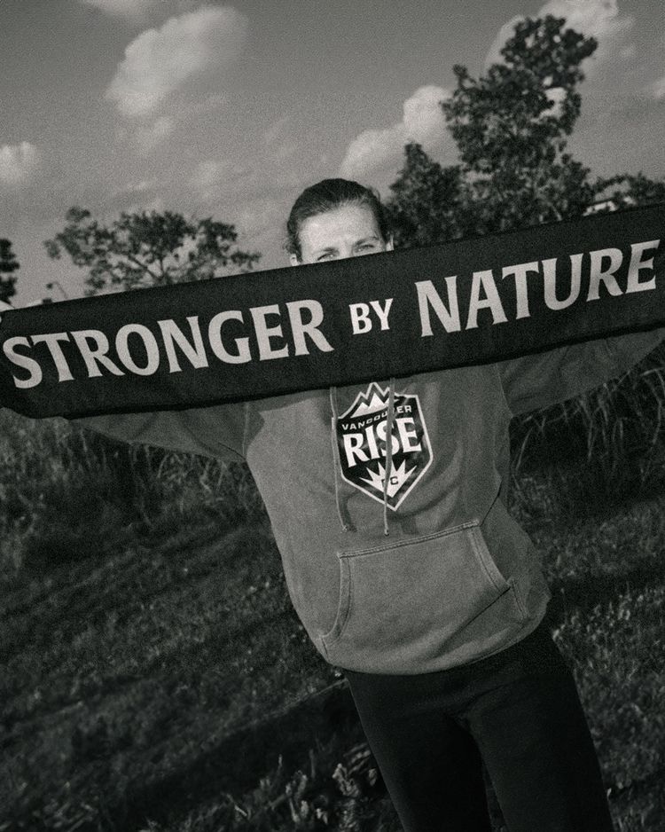 In black and white, Canadian soccer legend Christine Sinclair stands with an outstretched scarf covering her face. The scarf reads ‘stronger by nature.’