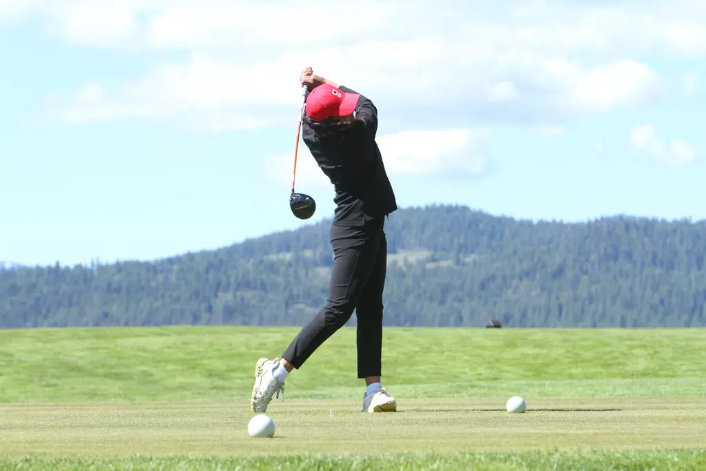 A girl with a red baseball cap, black pants, and a black long sleeve swings a driver.