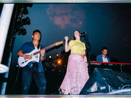 A woman in a yellow shirt and pink skirt stands by a microphone while two guys play a guitar (left) and set of keys (right) on each of her sides.