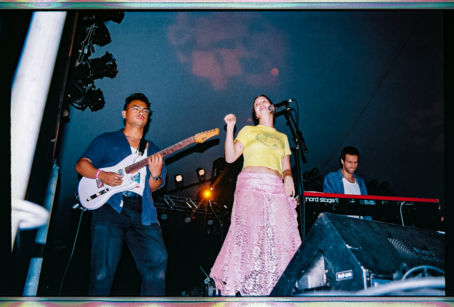 A woman in a yellow shirt and pink skirt stands by a microphone while two guys play a guitar (left) and set of keys (right) on each of her sides.