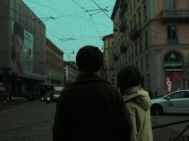 A dim, moody street in a city. Two people stand with their backs to the camera, close together as if holding hands, each wearing a jacket.