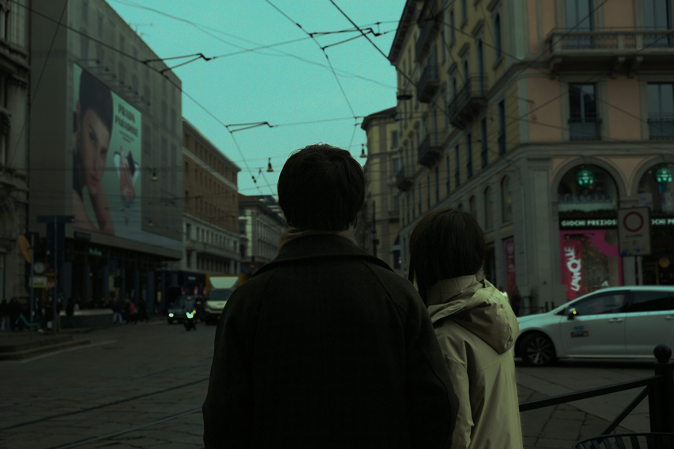 A dim, moody street in a city. Two people stand with their backs to the camera, close together as if holding hands, each wearing a jacket.