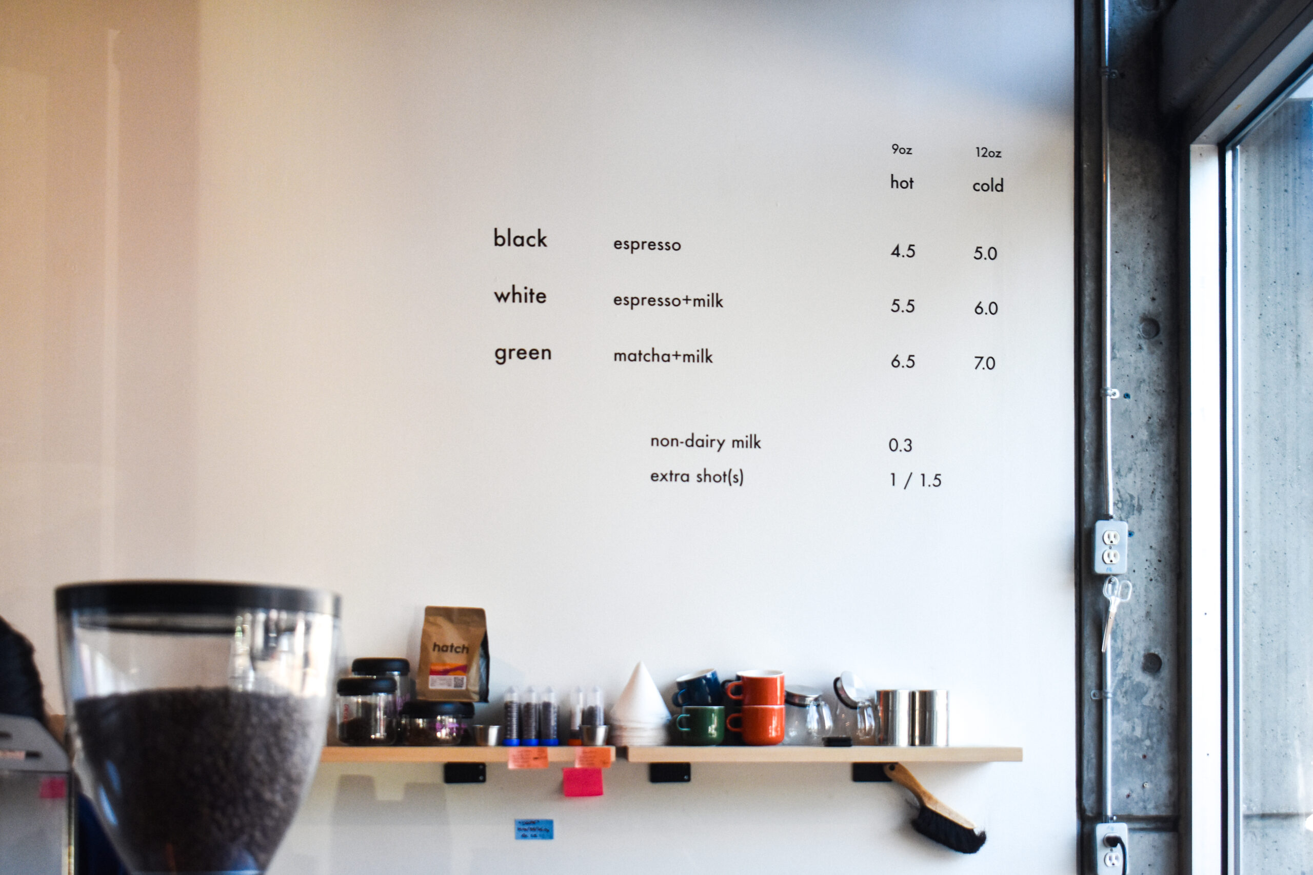 A plain white wall decorated with simple black writing that lists a menu that reads “black (espresso),” “white (espresso+milk),” and “green (matcha+milk).”