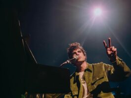 A man holds two fingers up as he sits at a piano with a spotlight shining in the back.