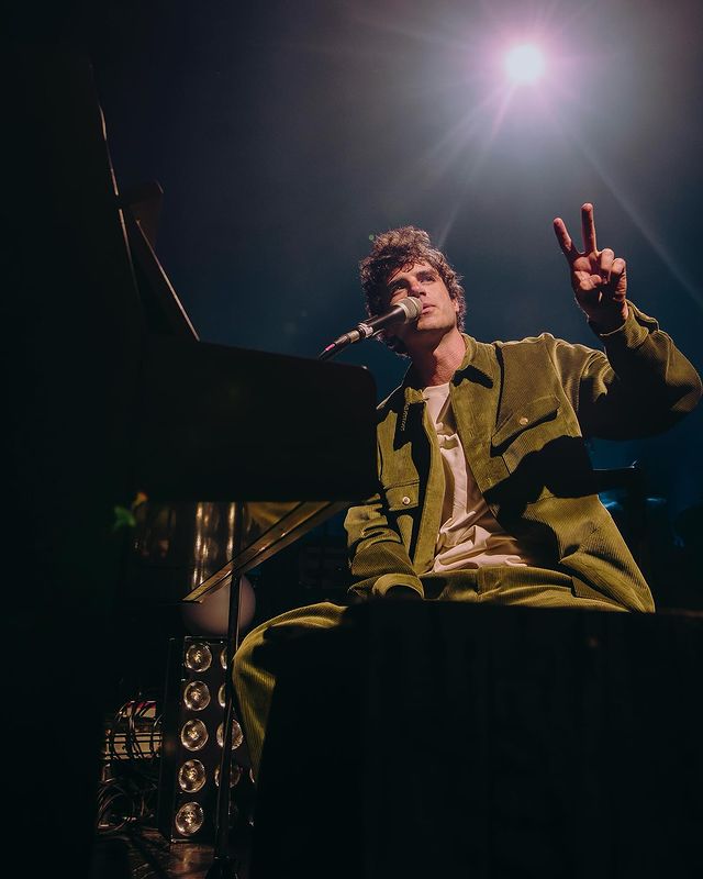 A man holds two fingers up as he sits at a piano with a spotlight shining in the back.