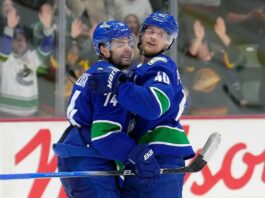 Two men in blue jerseys with a green stripe on the sleeve and blue helmets hug as they look to the side. Behind them are the boards and glass of a hockey rink. Cheering fans are visible through the glass.