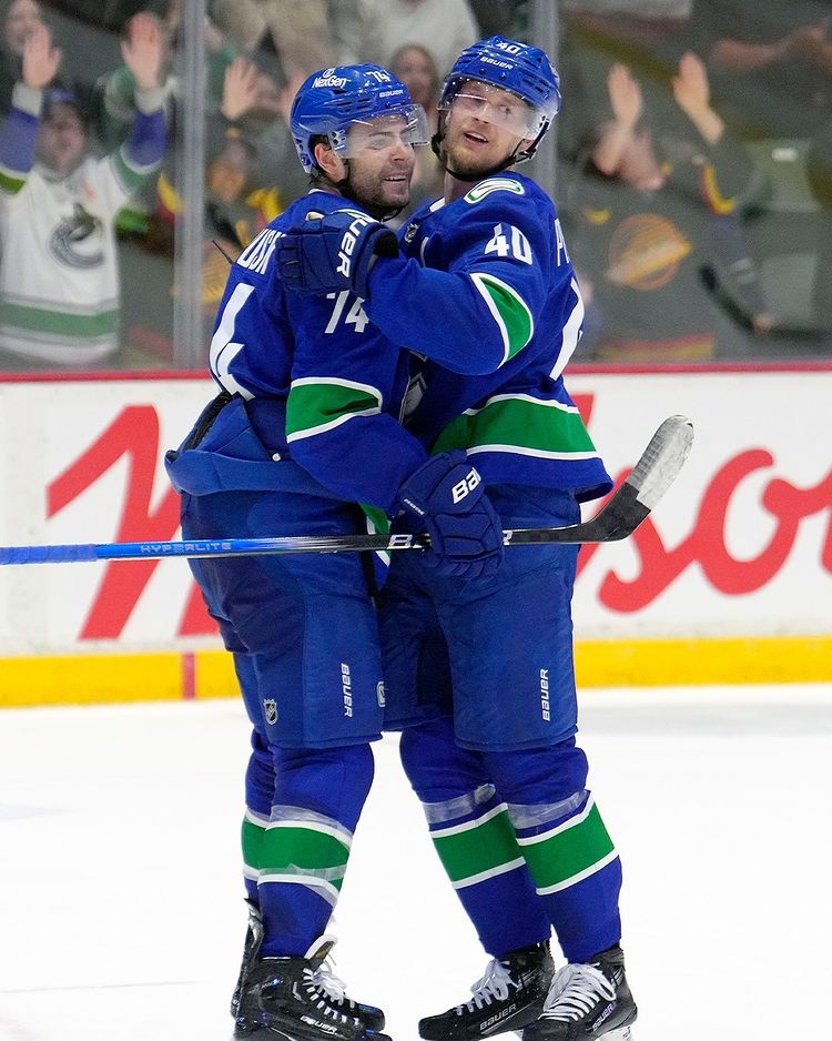 Two men in blue jerseys with a green stripe on the sleeve and blue helmets hug as they look to the side. Behind them are the boards and glass of a hockey rink. Cheering fans are visible through the glass.