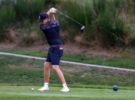 A man swings a golf club over his shoulder while wearing a black baseball cap decorated with an SFU logo.