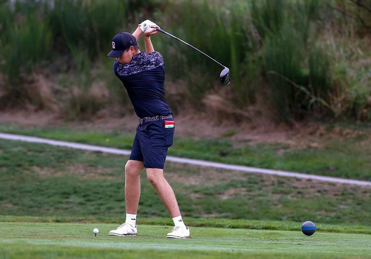 A man swings a golf club over his shoulder while wearing a black baseball cap decorated with an SFU logo.