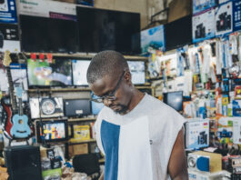 A man tinkers with an electronic device set up with outlets. Around him are various cords, wires, batteries, and other electronic supplies.