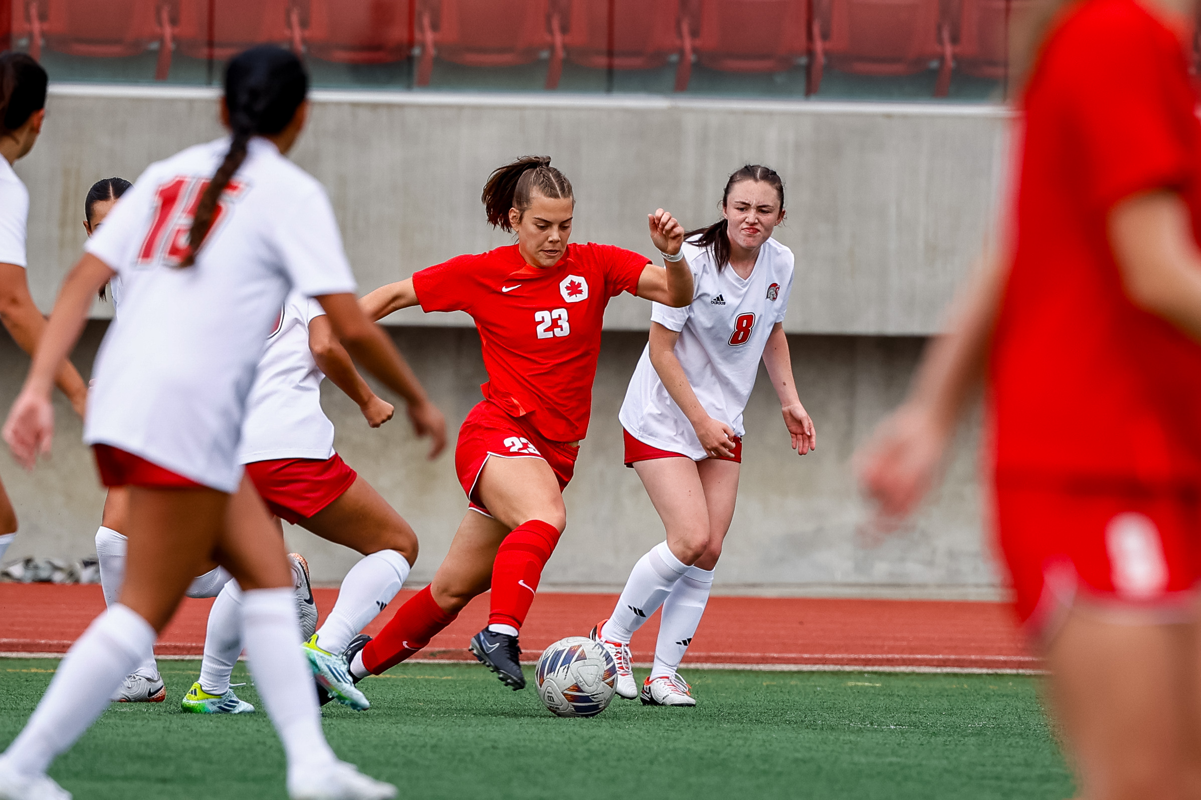 A player with a red uniform on and a maple leaf in the top corner kicks the ball around multiple players in white.