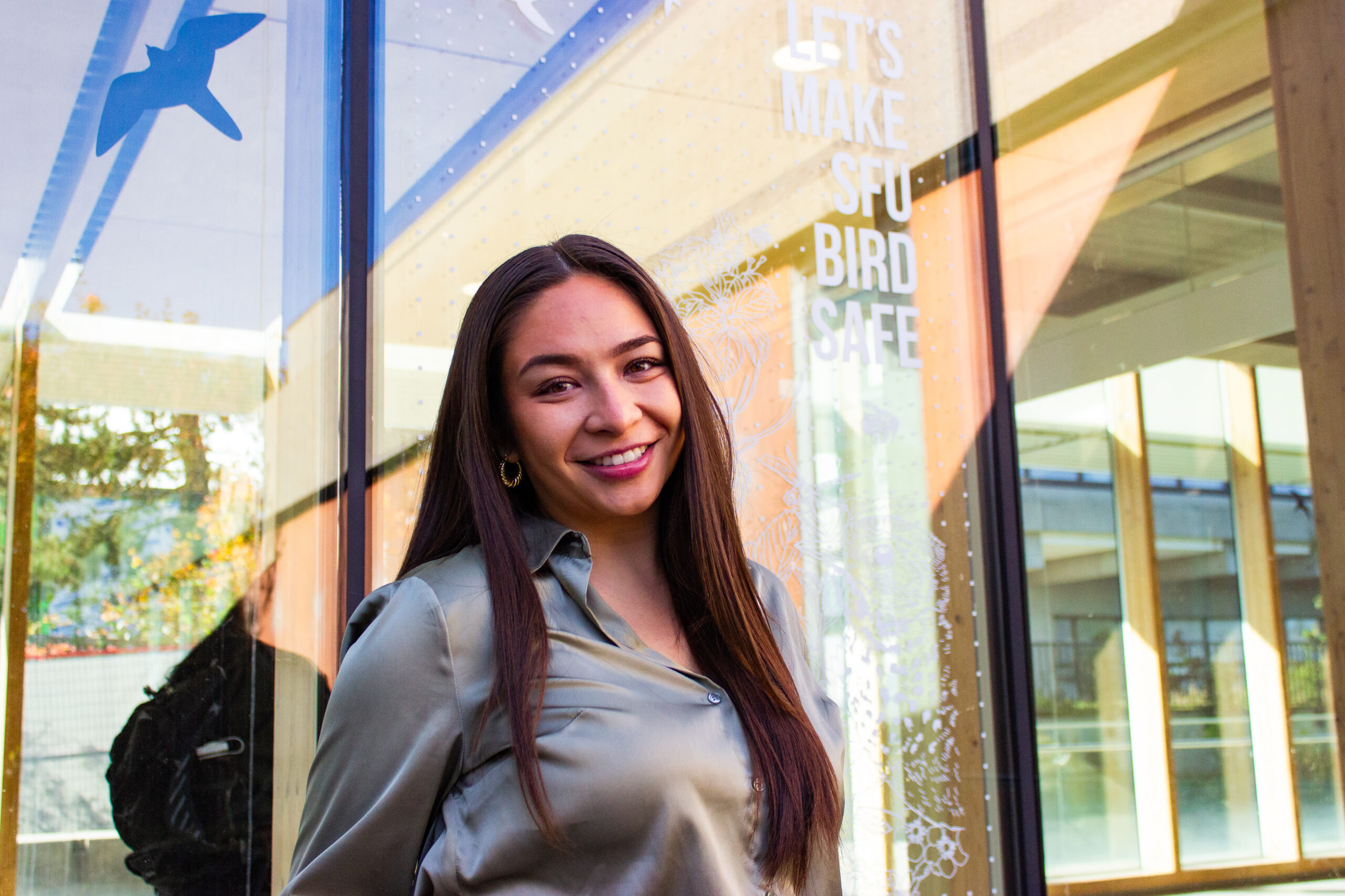 This is a photo of Vanessa Hum in front of a Feather Friendly dots window solution. It says, “LET’S MAKE SFU BIRD SAFE.”