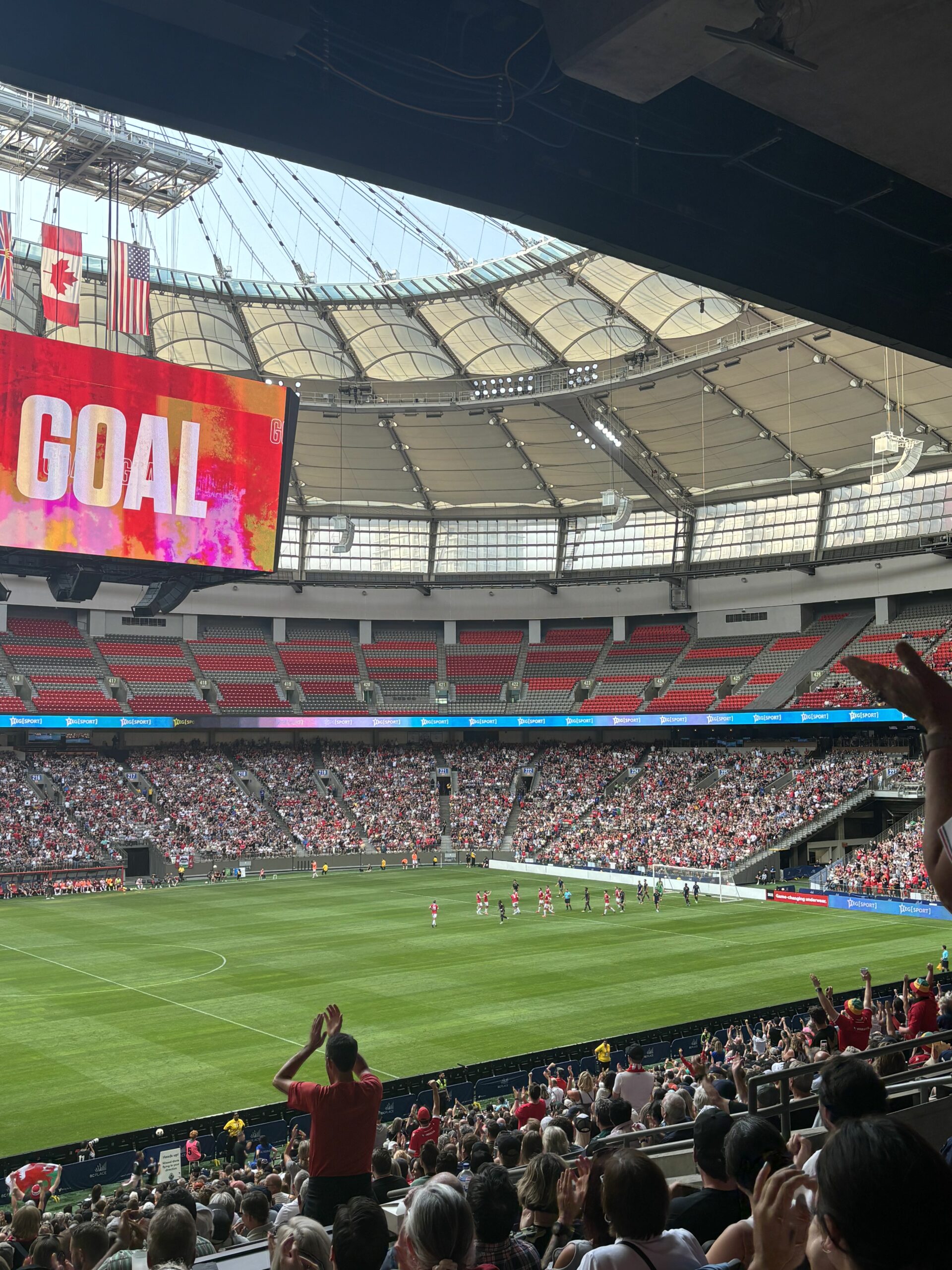 A large stadium decked out in red, white, and blue. A jumbotron hanging from the ceiling screens “GOAL” in white against a red background. Players gather on a grassy field.