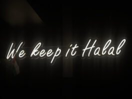 Against a dark background, a bright, white light-up sign reads ‘we keep it halal’ in cursive. Beside the sign is a Palestinian flag.