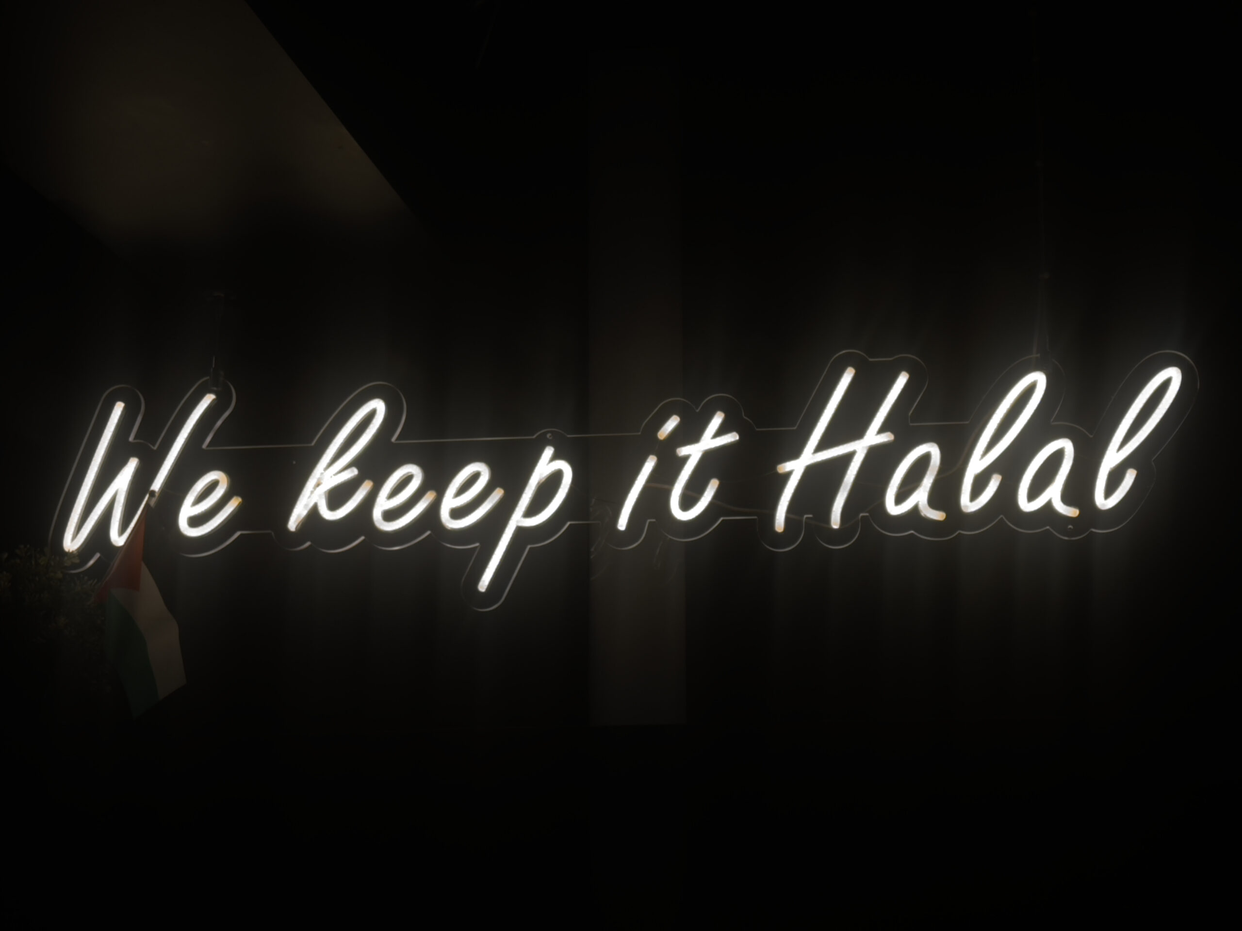 Against a dark background, a bright, white light-up sign reads ‘we keep it halal’ in cursive. Beside the sign is a Palestinian flag.