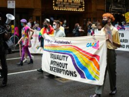 A group of masked protesters marching down a road. They are holding up a sign with a rainbow that says equality without exemptions.