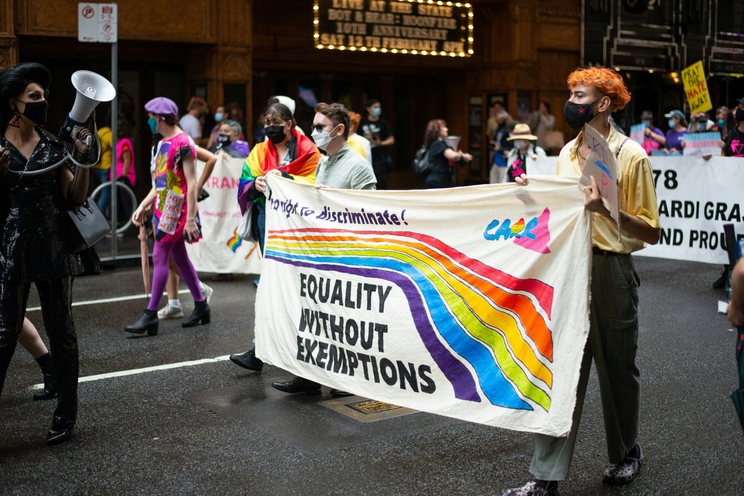 A group of masked protesters marching down a road. They are holding up a sign with a rainbow that says equality without exemptions.