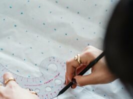 A person holds a ruler-labeled stencil down on a sheet of marked paper. Using another hand, they use a pen to trace around the stencil.