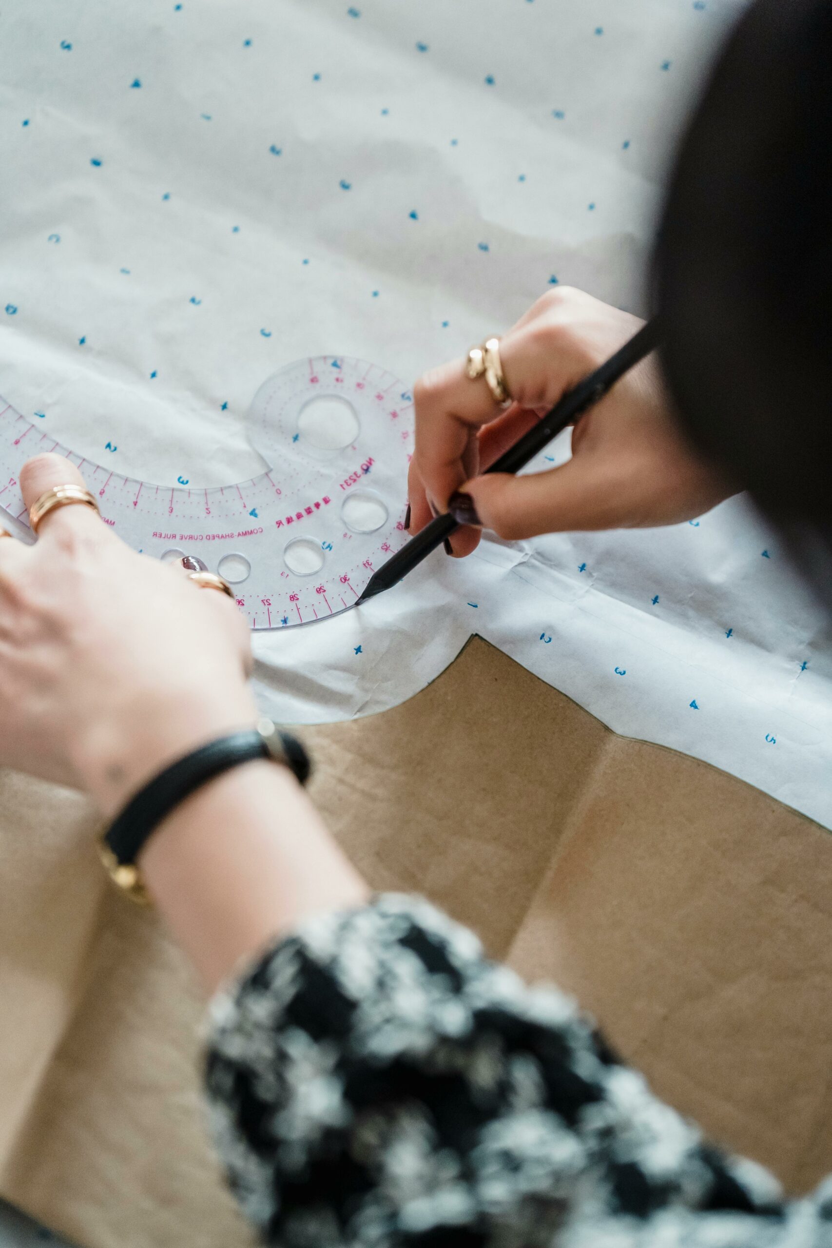 A person holds a ruler-labeled stencil down on a sheet of marked paper. Using another hand, they use a pen to trace around the stencil.