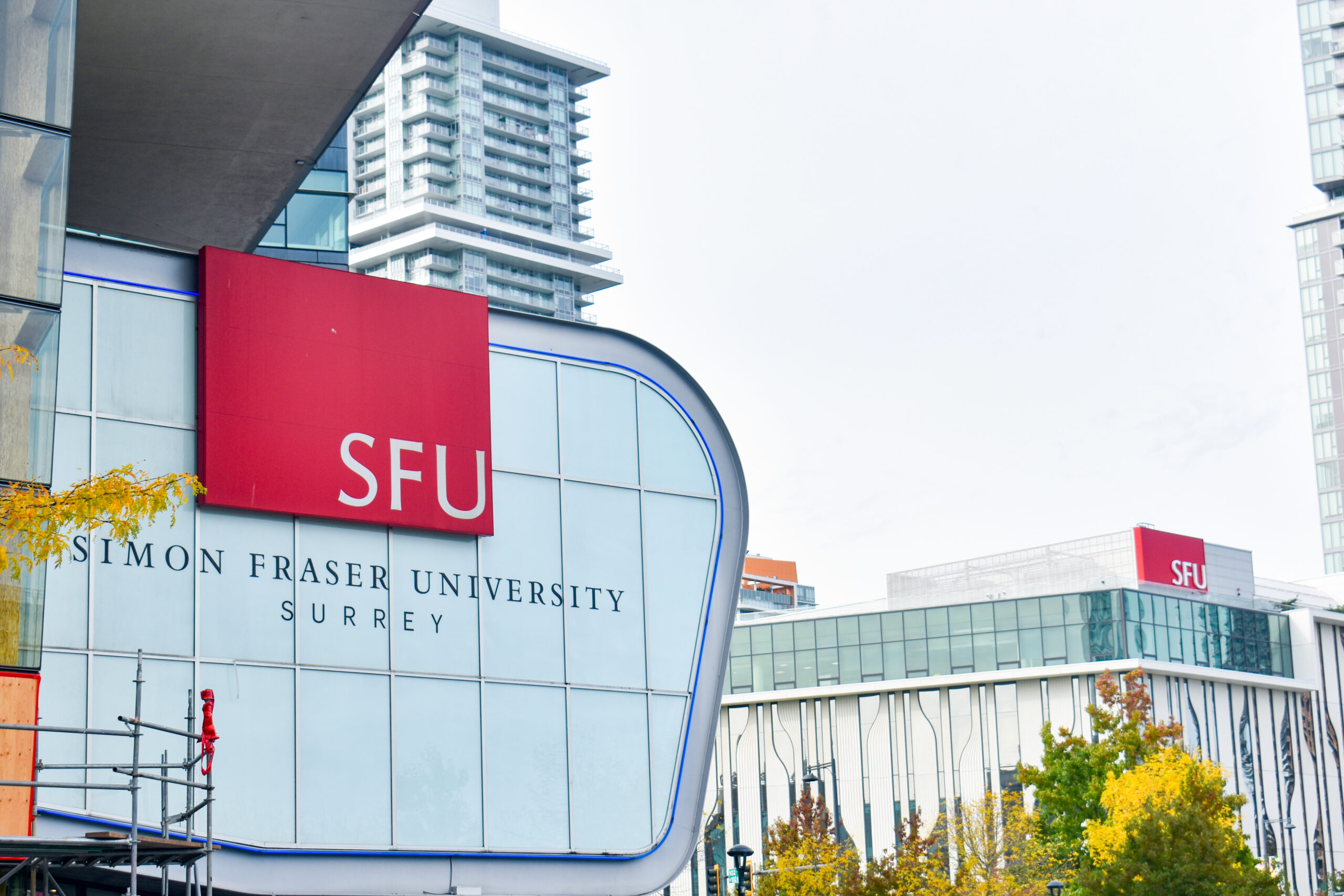 The side of a building holds a red sign reading ‘SFU’. Below it are the words “Simon Fraser University” and Surrey. The tops of tall condominiums are shown in the background.