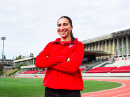 A girl with brown hair tied back smiles at the camera. She wears a red sweatshirt with “SFU” in the top right corner. Behind her is a set of bleachers.