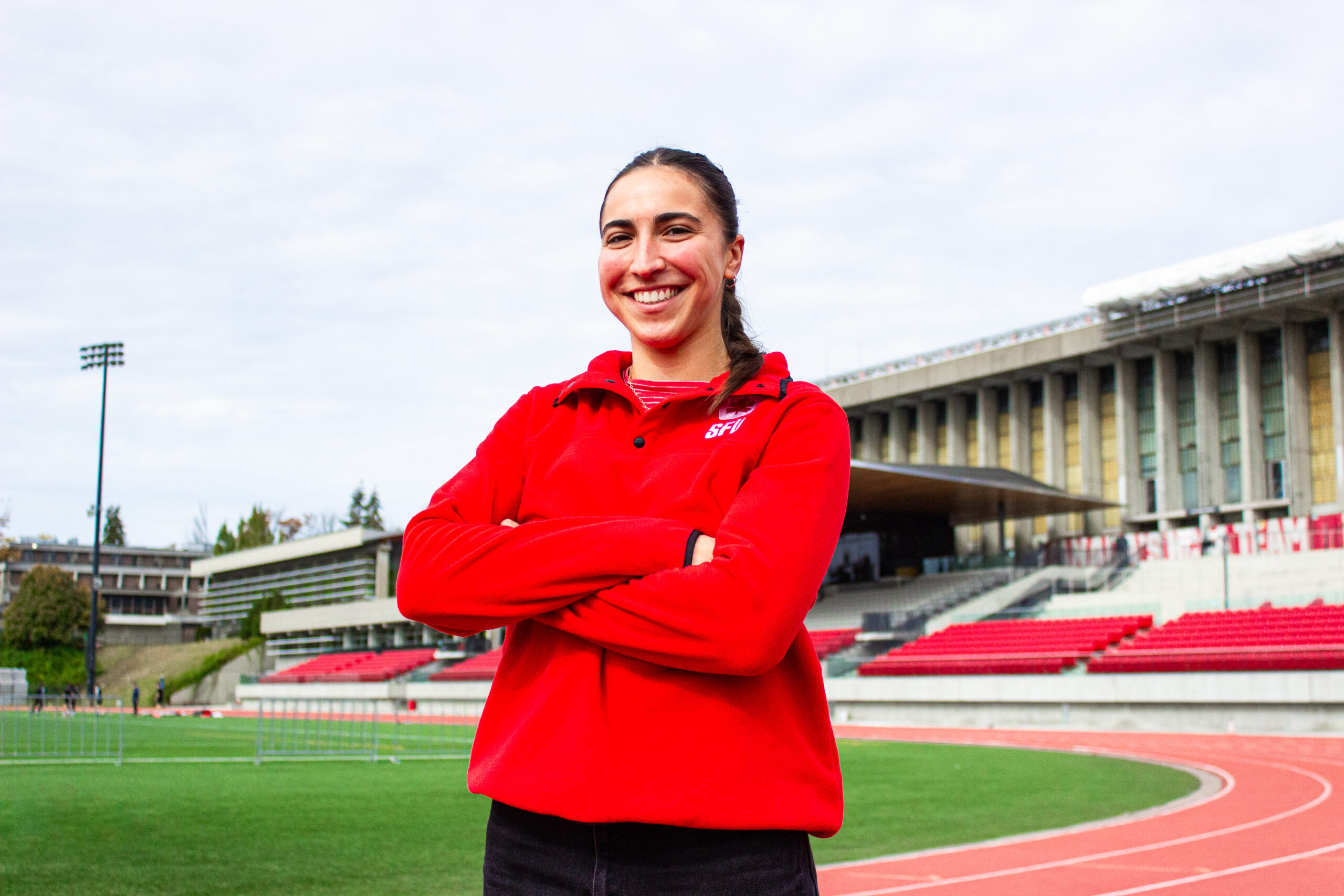 A girl with brown hair tied back smiles at the camera. She wears a red sweatshirt with “SFU” in the top right corner. Behind her is a set of bleachers.