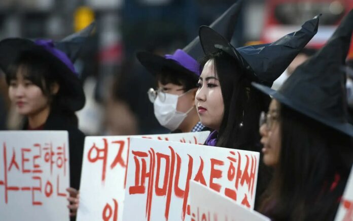 This is a photo of three people in witch hats holding up signs in Korean as part of the 4B movement.