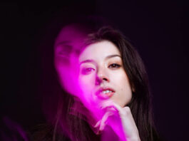 A woman with dark hair poses with her hand resting under her chin. Purple light streams off of her face with a shadowy effect.