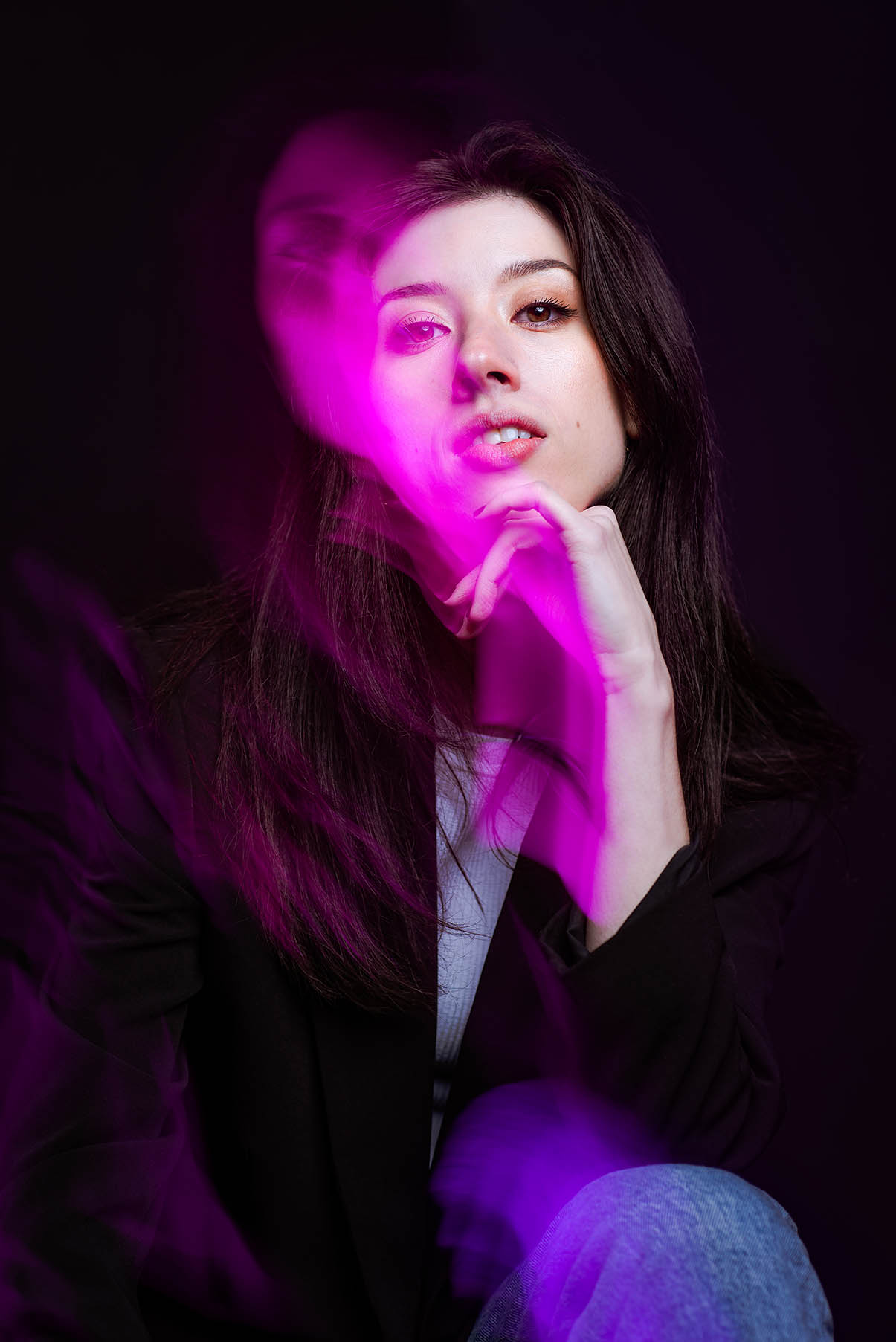 A woman with dark hair poses with her hand resting under her chin. Purple light streams off of her face with a shadowy effect.