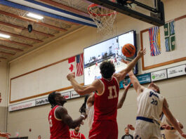 A man in a red basketball jersey jumps to swat a basketball away from a basketball hoop.