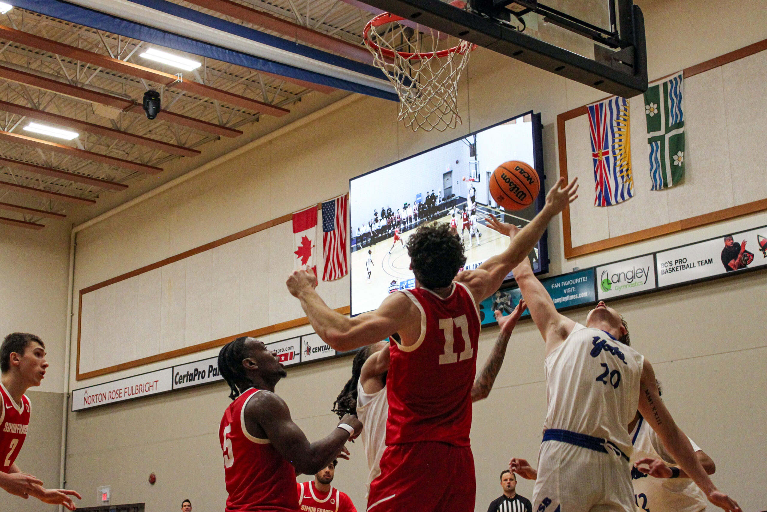 A man in a red basketball jersey jumps to swat a basketball away from a basketball hoop.