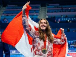 A woman in a Canadian Olympic uniform waves to a crowd behind the camera as she holds a Canadian flag on her shoulders.