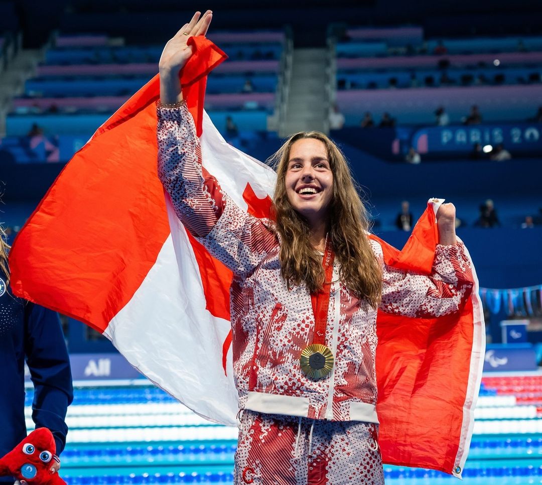 A woman in a Canadian Olympic uniform waves to a crowd behind the camera as she holds a Canadian flag on her shoulders.